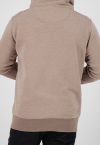 Uomo che indossa una felpa leggera color nocciola e pantaloni neri, visto da dietro su uno sfondo chiaro e semplice.