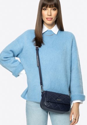 Žena s rovnými hnědými vlasy, oblečená v světle modrém svetru, bílé košili s límečkem, světlých džínách a nesoucí tmavě modrou prošívanou kabelku přes rameno.