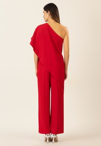Jumpsuit rosso asimmetrico con design drappeggiato e pantaloni a gamba larga. Realizzato in un tessuto morbido, abbinato a scarpe con tacco alto argentate.