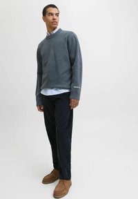 Pull en maille gris avec un col côtelé, porté sur une chemise blanche à collier. Un pantalon en velours côtelé foncé et des chaussures en daim marron complètent la tenue.