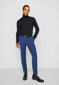 Marinefarbener Rollkragenpullover, enganliegende blaue Hose mit schmal zulaufendem Bein und schwarze Schuhe. Der Gürtel hat einen kontrastierenden blauen Akzent. Glatte Textur.