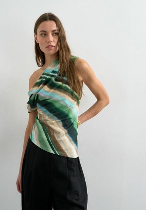 Femme aux longs cheveux bruns portant un haut à épaules dénudées avec des rayures vertes, bleues et beiges et un pantalon noir ample, devant un mur blanc uni.