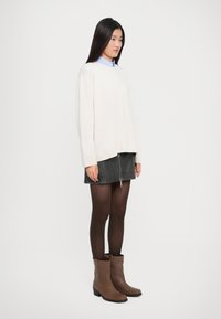 Giovane donna con lunghi capelli scuri che indossa un maglione bianco, una camicia blu con colletto, una gonna nera con zip frontale, collant neri e stivaletti alla caviglia marroni.