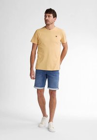 Keltainen puuvillainen t-paita pyöreällä kaula-aukolla ja pienellä logolla, yhdistetty farkkushortseihin, joissa on käännetyt reunat, ja valkoisiin tennareihin.