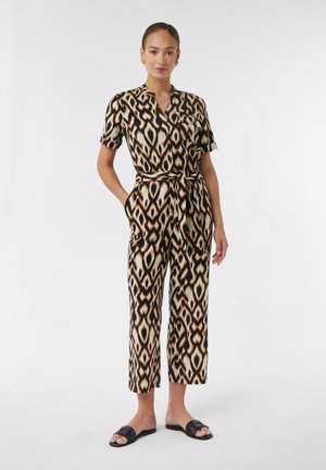 Braun- und cremefarbener Jumpsuit mit Ikat-Muster, kurzen Ärmeln und tailliertem Gürtel. Er verfügt über einen Kragen und Taschen. Schwarze Riemensandalen.