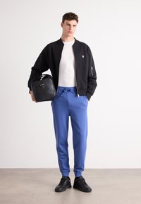 Zwarte bomberjack over een wit T-shirt, gecombineerd met blauwe joggingbroeken en zwarte sneakers. Inclusief een zwarte rugzak in de hand van het model.