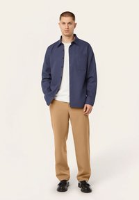 Marineblauwe button-up shirt met een zak, gecombineerd met beige broek. Persoon staat met handen in de zakken, met daaronder een wit T-shirt.