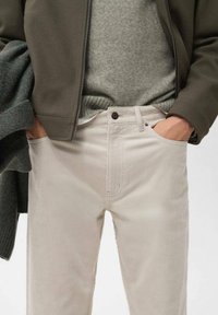 Pantaloni in corduroy beige chiaro con una vestibilità dritta, caratterizzati da una chiusura anteriore con bottone e tasche laterali, indossati con un maglione grigio e una giacca verde scuro.