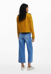Senfgelber, taillierter Mantel mit glatter Textur und Schulterklappen, kombiniert mit hellblauen, weit geschnittenen Jeans.