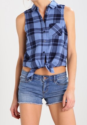 Ärmelloses blau-schwarz kariertes Hemd mit Knöpfen, am Taillenbund gebunden, kombiniert mit hellblauen Jeans-Shorts mit fransigem Saum.