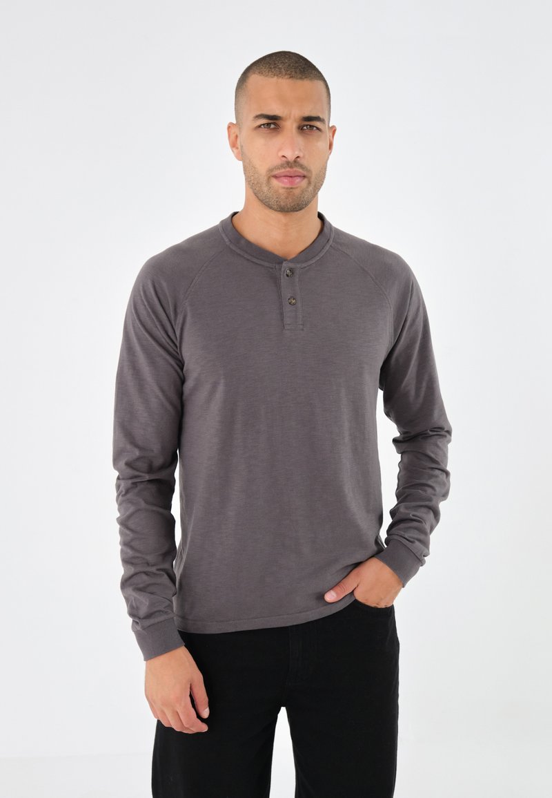 Langärmliges, dunkelgraues Henley-Shirt mit glatter Textur, Drei-Knopf-Blende und Raglanärmeln, kombiniert mit schwarzen Hosen.
