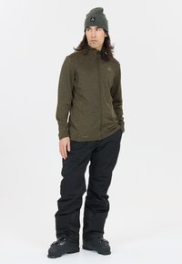 Olivengrøn zip-jakke med struktureret stof, parret med løse sorte snebukser og sorte skistøvler. Grå hue med logo-detalje.