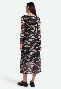 Langärmliges Kleid mit einem schwarz-beigen abstrakten Muster, transparenten Ärmeln, tailliertem Schnitt und einem fließenden, knöchellangen Rock. Schwarze Schuhe runden den Look ab.