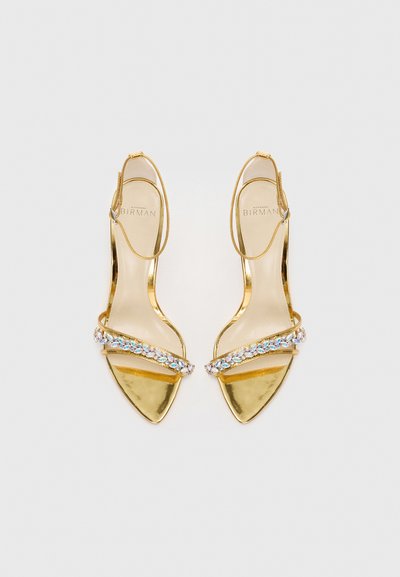 Sandali con tacco alto dorati, punta a punta, cinturino decorato con pietre trasparenti e chiusura alla caviglia. Finitura metallica liscia con un design elegante e minimalista.