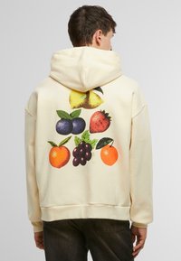 Cremefarvet hættetrøje med et grafisk design af forskellige frugter: gul æble, blå blomme, rød jordbær, appelsin og lilla drue med grønne blade.
