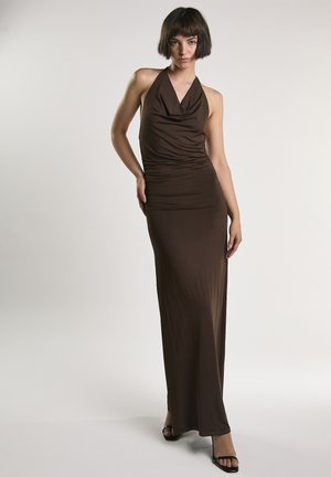 Kobieta o krótkich włosach ubrana w dopasowaną, bezrękawnikową, ciemnobrązową suknię maxi na szyję oraz czarne sandały na obcasie z odkrytymi palcami, stojąca na jednolitym jasnym tle.