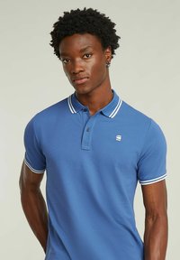 Blå polo skjorta i bomull med en tvåknappsslutning, vita accentremsor på kragen och ärmarna samt en liten logotyp på bröstet.