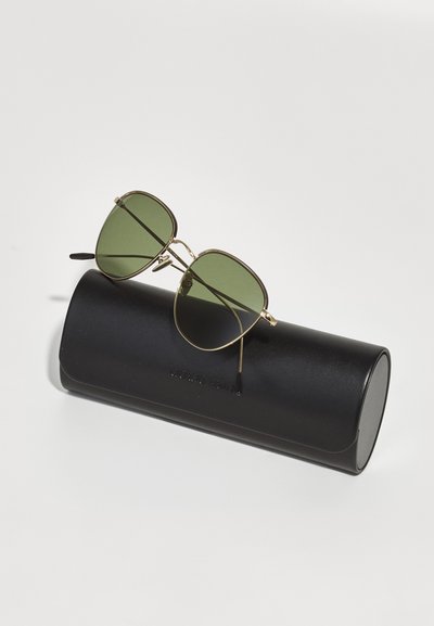 Sunglasses med guldinramning och gröna linser som vilar på ett svart cylindriskt etui. Ytan på etuiet är slät med präglad text.