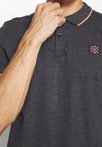 Homme ajustant le col d'un polo gris foncé avec des finitions corail et un petit écusson sur la poitrine portant l'inscription "1962 T T."