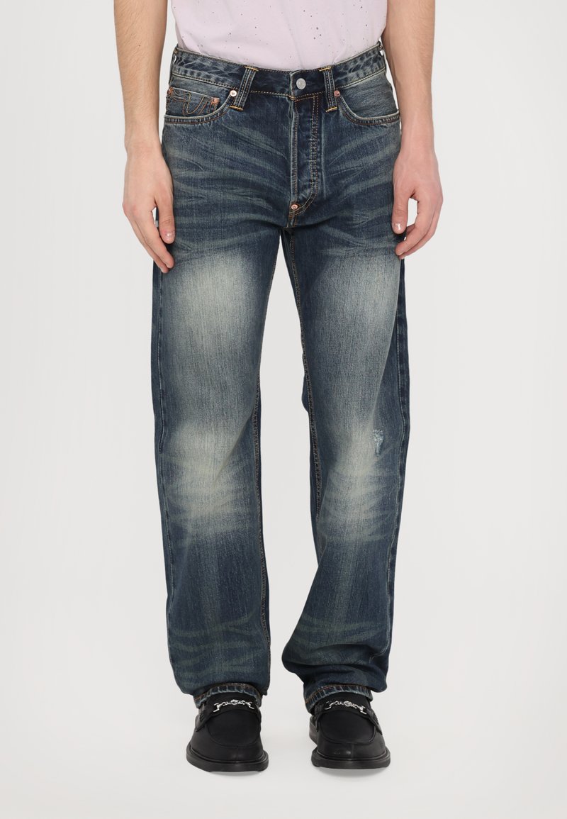 Jean en denim bleu foncé avec une coupe décontractée, légères décolorations et détails usés. Dispose d'une fermeture à bouton et d'accents de couture orange.