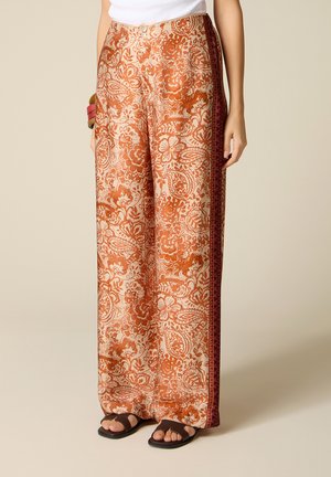 Donna con pantaloni a gamba larga con motivo floreale paisley arancione e strisce laterali rosse, abbinati a sandali neri e top bianco.