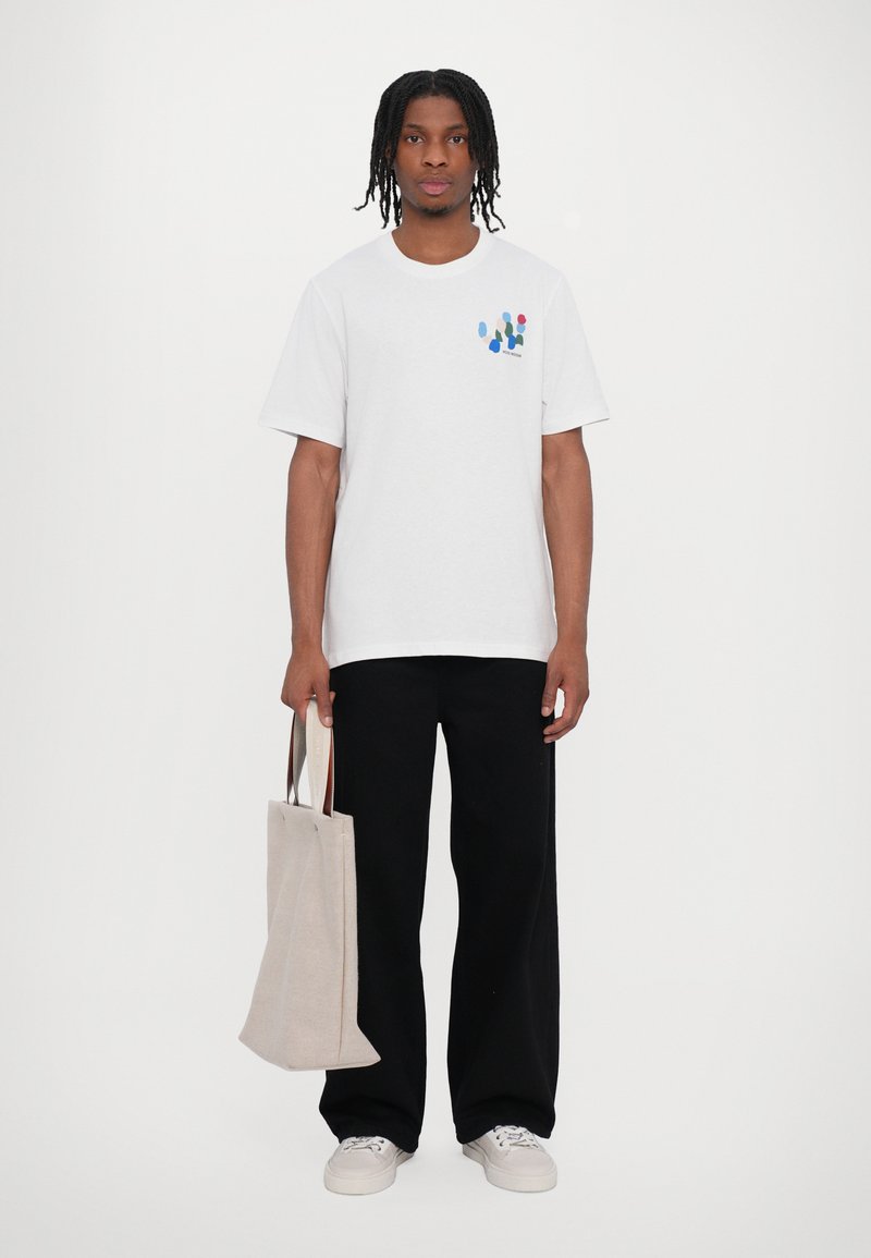 White cotton t-shirt with abstract multi-colored print on left chest, paired with black loose-fit pants and a beige canvas tote bag.