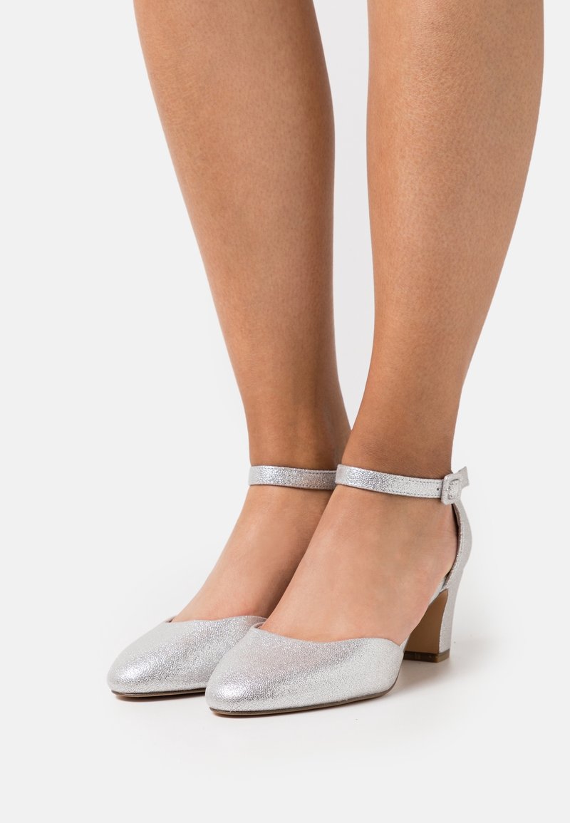 Jambes portant des chaussures argentées à petit talon bloc, bouts fermés pointus et brides à la cheville sur un fond clair uni.
