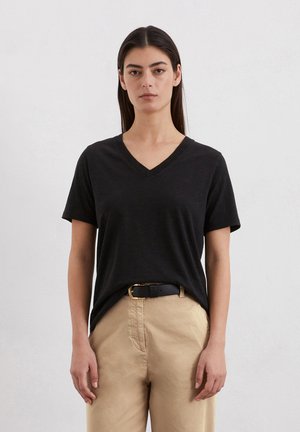 T-shirt noir à col en V en tissu doux, à manches courtes, associé à un pantalon beiges et une ceinture noire avec une boucle dorée.