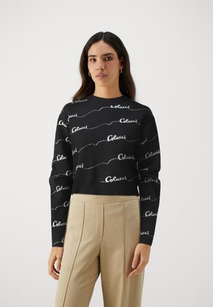 Sort sweater med hvid skriftlogo-mønster, ribbede ærmer og kant, rundt halsudskæring, parret med lysebeige højtaljede bukser.