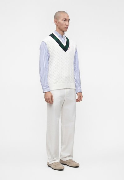 Homme debout portant un gilet en maille torsadée blanc avec une bordure verte, une chemise rayée bleue, un pantalon blanc et des mocassins beige sur un fond uni.