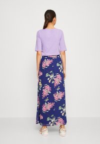 Kvinna med mörkt hår i hästsvans bär en ljuslila topp, lång marinblå blommig kjol och vita sneakers, stående vänd mot en enfärgad vit vägg.