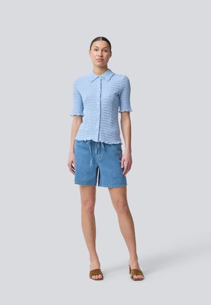Camisa azul clara de manga corta con tejido texturado y cuello, combinada con shorts de mezclilla azules y sandalias de tacón marrones.