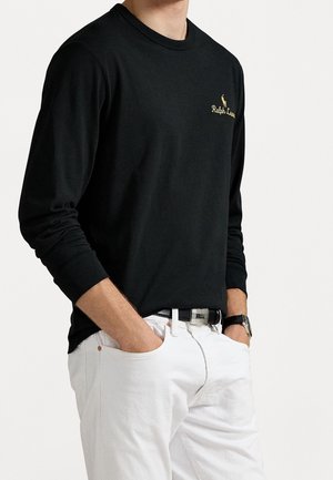 Mies, jolla on musta pitkähihaisen Ralph Lauren -paita, jossa logo rinnassa, valkoiset housut, musta vyö ja rannekello, kädet taskuissa.