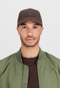 Bărbat purtând o șapcă maro Alpha Industries, o geacă bomber verde și o cămașă maro, așezat cu fața spre cameră pe un fundal simplu, deschis.
