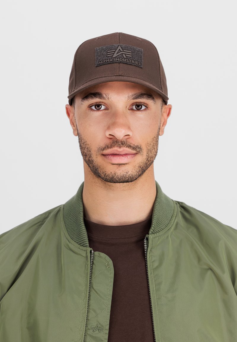 Bărbat purtând o șapcă maro Alpha Industries, o geacă bomber verde și o cămașă maro, așezat cu fața spre cameră pe un fundal simplu, deschis.