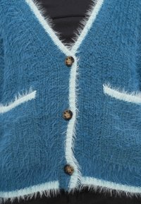 Μπλε χνουδωτό ζακέτα με λευκή επένδυση, διαθέτει τρία καφέ κουμπιά, δύο μπροστινές τσέπες και σχέδιο με λαιμόκοψη V. Ευδιάκριτη μαλακή υφή.