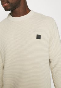 Lys beige strikkegenser med rund hals, med et teksturert ribbemønster og et lite svart logo-patch på brystet.