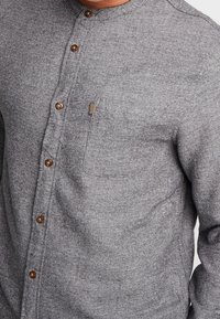 Camisa de botones gris hecha de tela texturizada, con un diseño sin cuello, botones marrones y un pequeño bolsillo en el pecho con detalles de acento.