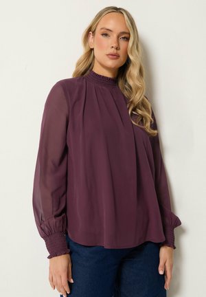 Vrouw met lang blond haar, die een paarse blouse met hoge hals en lange mouwen en donkerblauwe spijkerbroek draagt, staand tegen een lichte achtergrond.