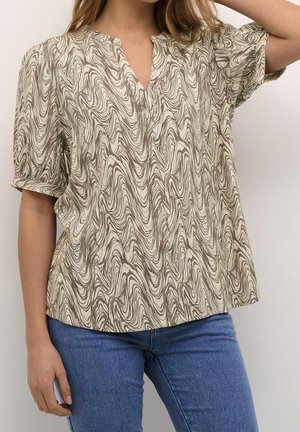 Femme portant un chemisier beige à manches courtes avec un motif abstrait ondulé marron et un jean bleu, un bras levé derrière la tête.