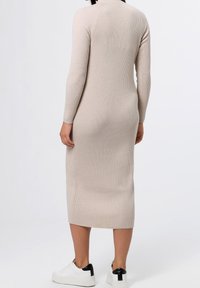 Abito beige lavorato a maglia con texture a coste, maniche lunghe e lunghezza midi. Indossato con scarpe bianche con un accento nero sul tallone.
