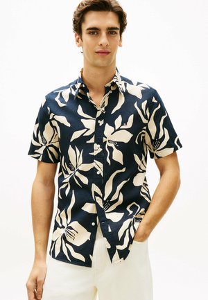 Jeune homme portant une chemise bleu marine à manches courtes avec de grands motifs floraux beige et un pantalon blanc, main dans la poche.