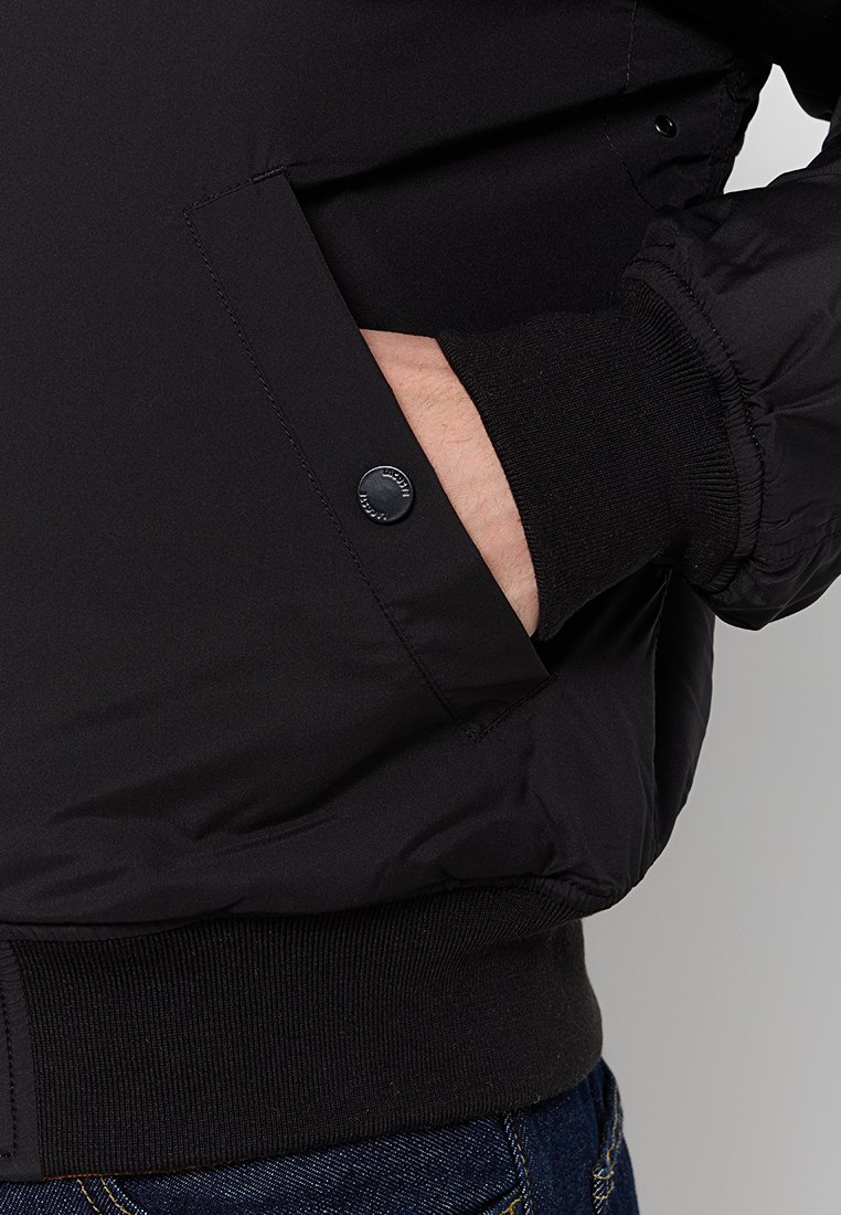 Veste noire avec un matériau extérieur lisse, dotée d'une poche à bouton-pression et de poignets côtelés. Mettez l'accent sur les détails de la poche et la texture.