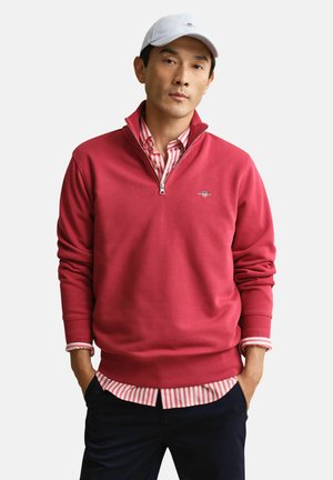 Mann trägt weiße Baseballkappe, rotes Viertel-Reißverschluss-Sweatshirt über einem rot-weiß gestreiften Hemd mit Knöpfen und dunkle Hose, steht mit den Händen in den Taschen.