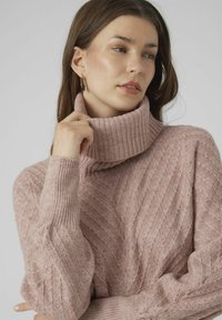 Light pink knitted sweater with a chunky ribbed turtleneck, featuring a chevron pattern and ribbed cuffs. Soft texture and relaxed fit.