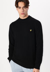 Maglione in maglia a coste nero con colletto alto, caratterizzato da un piccolo logo dorato di un'aquila sul petto. Texture morbida e maniche lunghe.