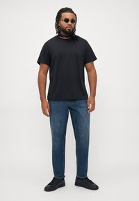 Man with dreadlocks wearing black t-shirt, blue jeans, black sneakers, sunglasses, and a black neck scarf standing against a plain light background.