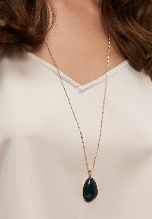 Goldkette mit ovalem türkisfarbenem Anhänger, glatte Oberfläche, auf Brusthöhe hängend, getragen über einem hellbeigen V-Ausschnitt-Top.