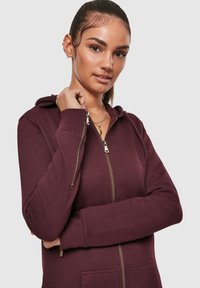 Temno rjava jopa z zadrgo, mehka na otip, z zlatimi kovinskimi dodatki, kapuco in rebrastimi manšetami. Prikaz bližnjega posnetka sprednjega dela in položaja rok.