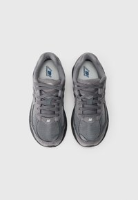 Pelēki sporta apavi ar sietveida augšdaļu, zamšādas akcentiem, plakanām šņorēm un tumšu gumijas zoli. New Balance logo ir redzams sānos.